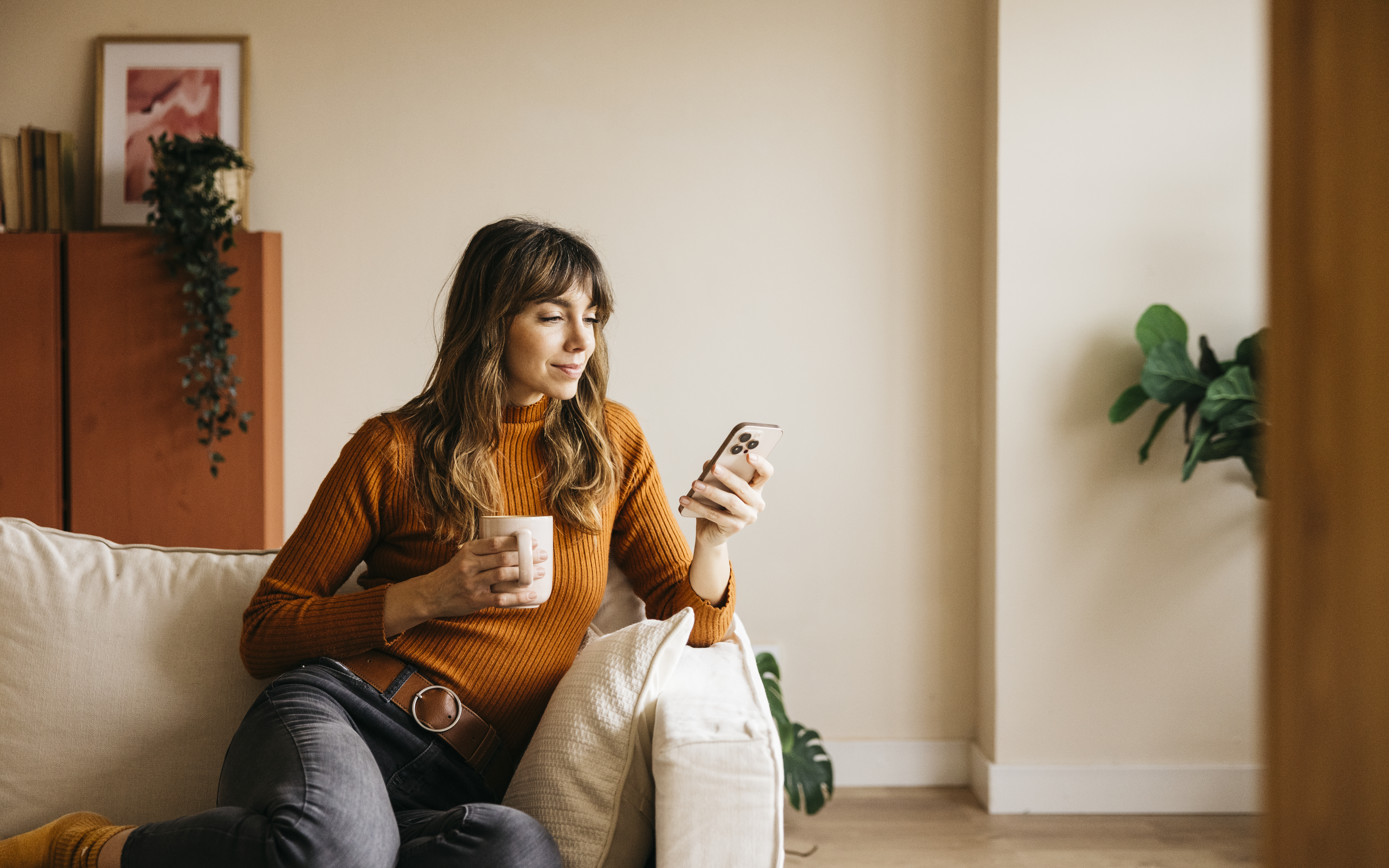Woman relaxing on a sofa, drinking from a mug and using her phone
